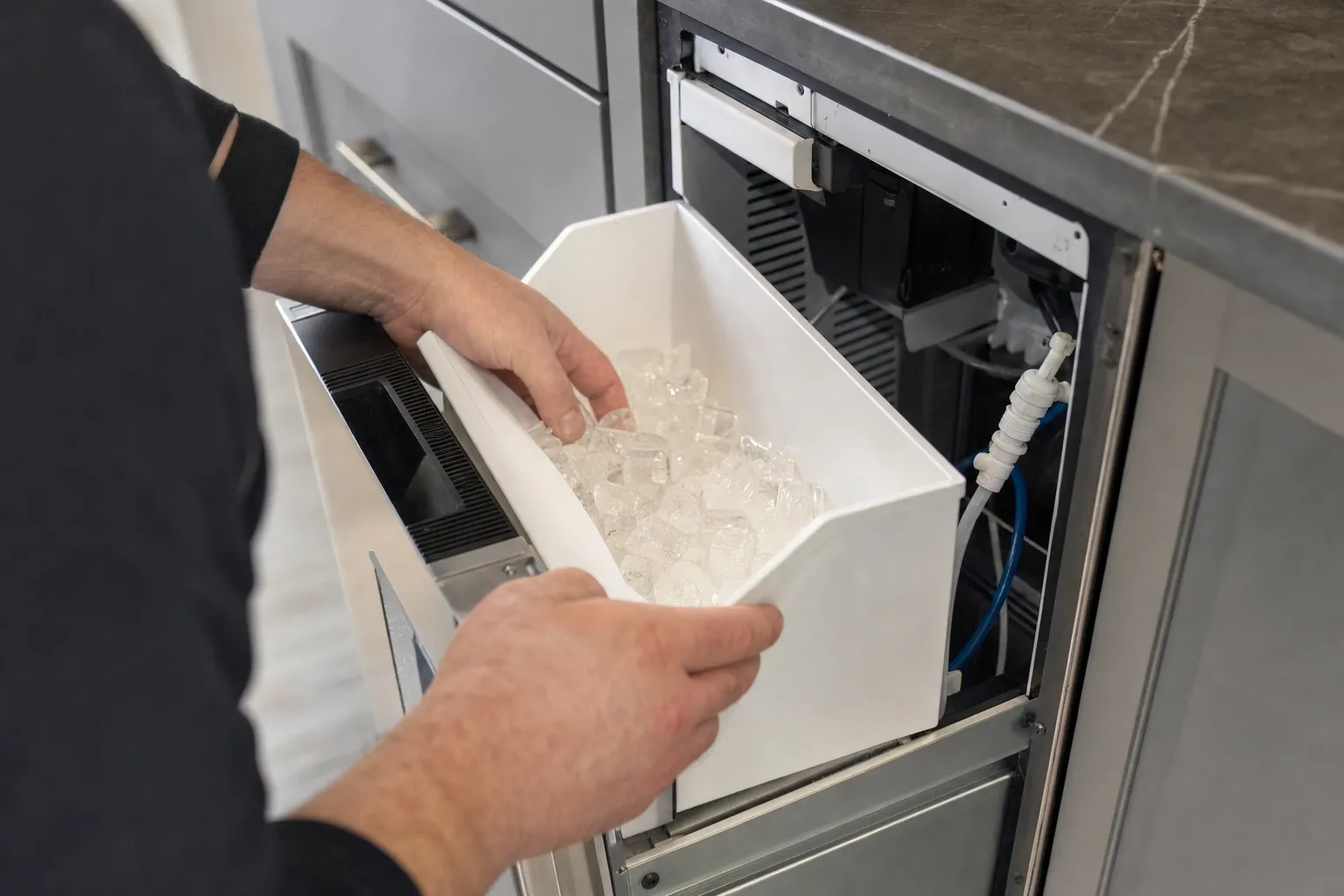 Hands lifting the ice storage bin out of an undercounter ice maker, water line visible behind