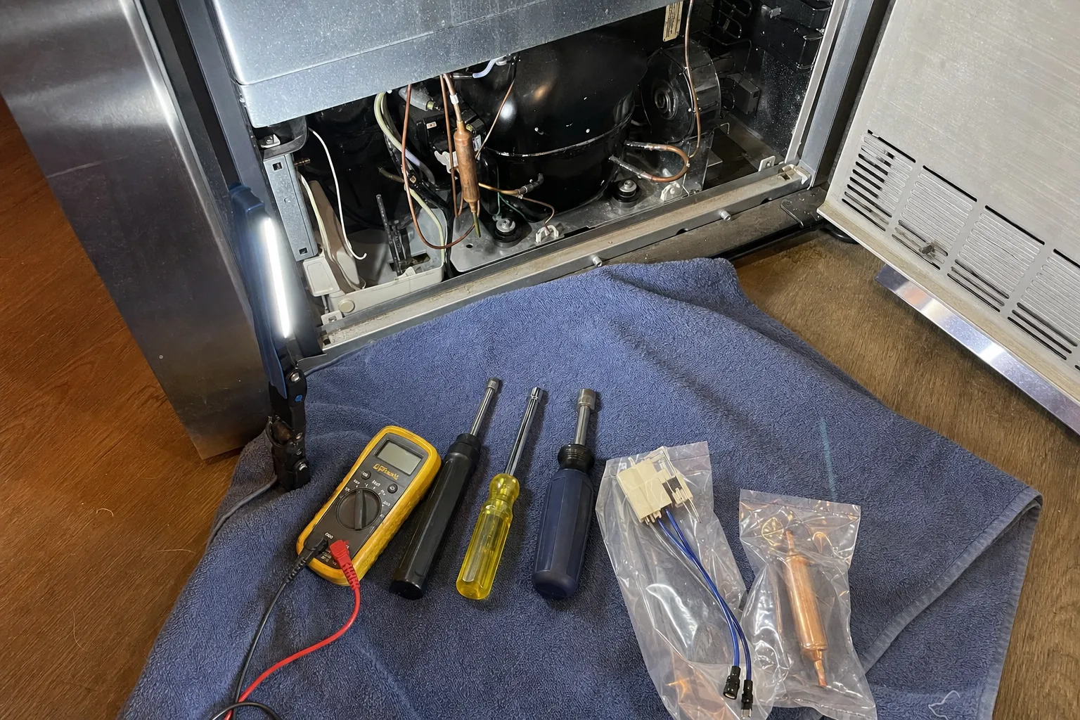 Multimeter, three nut drivers and two bagged replacement parts laid on a towel in front of an open compressor bay